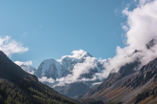 Foto paisaje escénico con bosque en otoño valle de montaña contra grandes montañas de nieve en nubes bajas en la luz del sol de la mañana abetos en la ladera con vistas a la alta cordillera nevada iluminada por el sol en nubes bajas