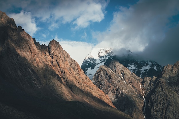 Paisaje escénico con grandes rocas y montañas nevadas a la luz del sol en nubes bajas. Maravillosa vista a la cima de la montaña con nieve bajo el sol en el cielo nublado. Un paisaje impresionante con un pináculo blanco como la nieve iluminado por el sol.