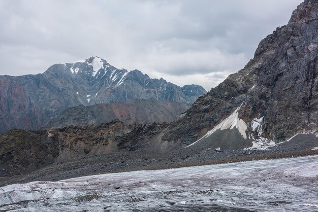 Paisaje impresionante con una larga lengua de glaciar contra una alta cordillera con un pico rocoso afilado en un clima nublado Pico puntiagudo en forma de cono en un cielo nublado dramático Gran glaciar entre rocas afiladas