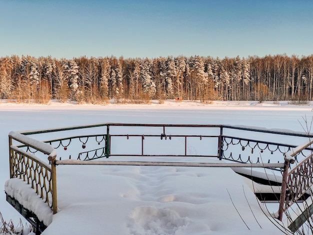 Paisaje invernal con antiguo muelle cubierto de nieve en la orilla del lago congelado con árboles forestales en horison