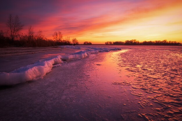 Paisaje invernal con río congelado y cielo ardiente al atardecer.