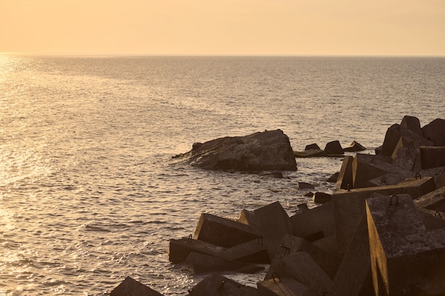 Paisaje marino con grandes rompeolas de hormigón en la hermosa puesta de sol naranja, olas del mar rompiendo en rompeolas hechos de rocas en el sol vespertino. Bloques y piedras para proteger la costa de tormentas