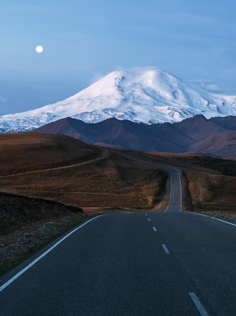 Paisaje matutino vertical con carretera sinuosa al Monte Elbrus bajo el cielo matutino con luna llena El camino vacío desaparece en la distancia en el crepúsculo Camino con vista en perspectiva marcada
