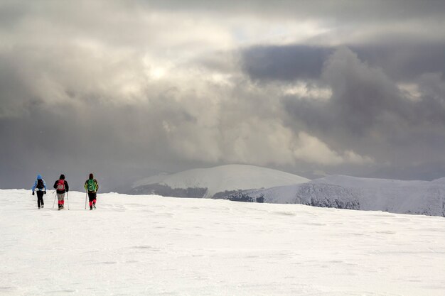 Paisaje de montaña de invierno. Tres viajeros excursionistas turísticos en ropa brillante con mochilas en campo nevado caminando hacia la montaña distante sobre fondo de espacio de copia de cielo azul oscuro nublado.