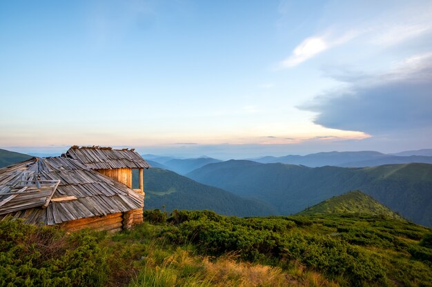 Paisaje de montaña de noche de verano con antiguo refugio turístico abandonado en colinas cubiertas de hierba y picos distantes al atardecer colorido.