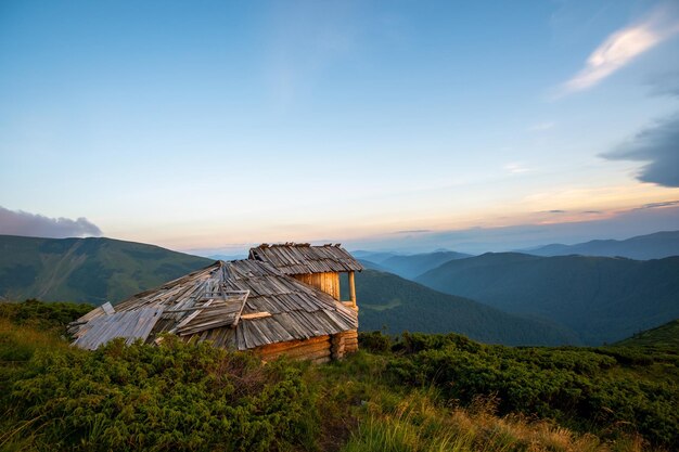 Paisaje de montaña de noche de verano con antiguo refugio turístico abandonado en colinas cubiertas de hierba y picos distantes al atardecer colorido.