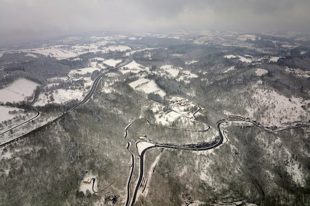 Paisaje neblinoso aéreo con carretera de montaña cubierta de nieve fresca caída durante fuertes nevadas en el bosque de montaña de invierno en un día frío y tranquilo