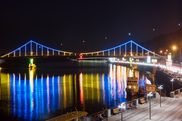 Paisaje nocturno. La ciudad de Kiev, Ucrania, Europa. Puente peatonal sobre el río Dnieper. Hermosa iluminación y reflejo en el agua en colores azul y amarillo.