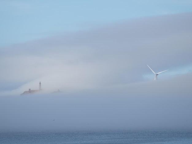 Paisaje entre nubes bajas donde se puede ver un faro y un molino de viento.