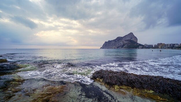 Paisaje panorámico en el mar con una gran montaña en el horizonte y olas rompiendo contra las rocas.