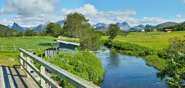 Paisaje de un río entre colinas y montañas Follaje verde junto a la orilla del río con un cielo azul en Noruega Aguas tranquilas cerca de un desierto vibrante contra un horizonte nublado brillante Escena de naturaleza pacífica