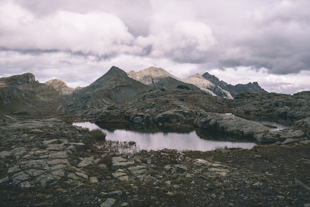 Paisaje rocoso de gran altitud y pequeño lago. Majestuoso paisaje alpino con espectacular cielo tormentoso. Visión gran angular desde arriba, imagen en tonos, filtro vintage, tonos divididos.