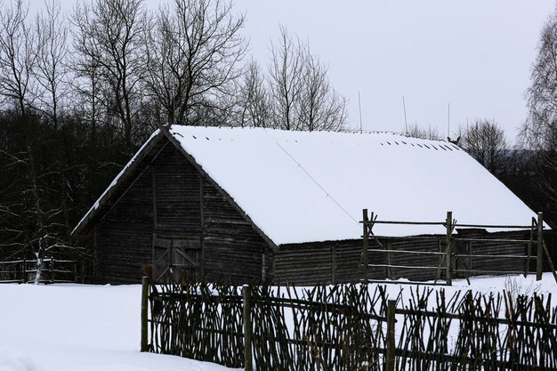 Paisaje ruso de invierno Una vieja cabaña de madera una casa de troncos con techo de paja Aldea rusa abandonada cubierta de nieve Casa de troncos con un granero con una cerca de mimbre de madera