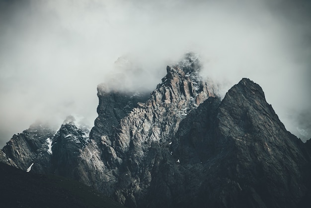 Paisaje surrealista atmosférico oscuro con la cima de la montaña rocosa oscura en nubes bajas en el cielo nublado gris. Nube gris baja en el pináculo alto. Roca negra alta con nieve en nubes bajas. Montañas sombrías surrealistas.