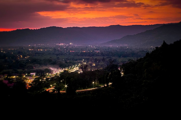 Paisaje con valle de montaña alpino nubes bajas bosque cielo púrpura con estrellas iluminación de la ciudad al atardecer Aéreo