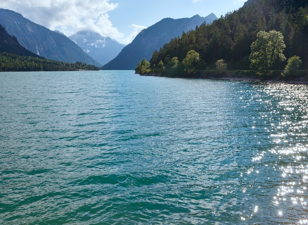 Paisaje de verano de Plansee con nieve en la ladera de la montaña y el sol brilla sobre la superficie del agua (Austria)