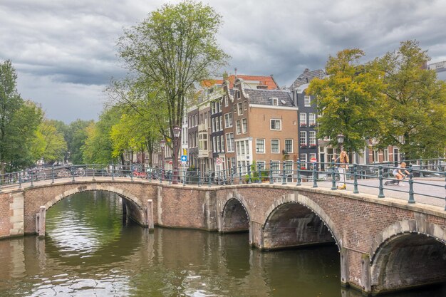 Países Bajos Día nublado de verano Antiguos puentes de piedra en los canales de Ámsterdam Casas típicas holandesas con fachadas danzantes en el terraplén Adolescentes en scooters