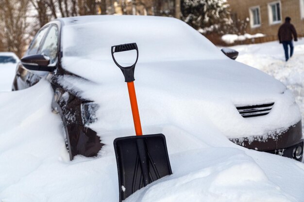 Pala de nieve de plástico delante de un coche cubierto de nieve en la soleada mañana de invierno