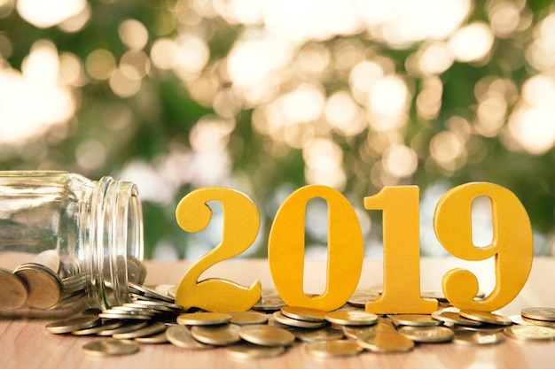 La palabra 2019 puso monedas y las botellas de cristal con las monedas dentro en fondo verde del bokeh.