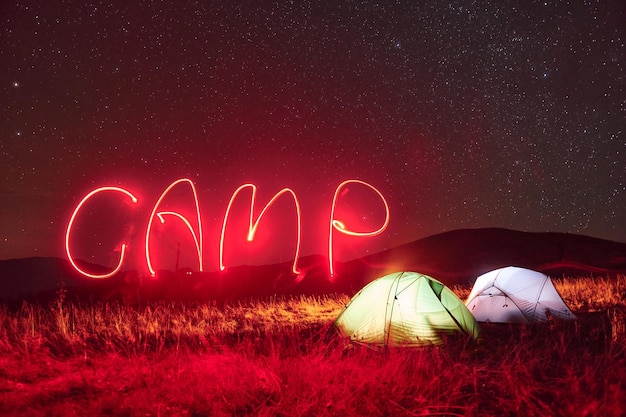 Palabra de campamento de neón rojo Dos carpas iluminadas bajo estrellas en las montañas por la noche