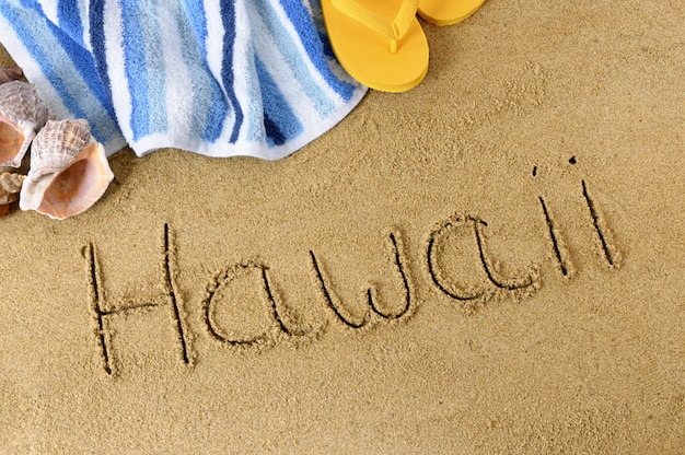 La palabra Hawaii escrita en arena con chanclas y toalla de playa