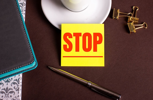 La palabra STOP escrita en papel amarillo sobre un fondo marrón cerca de una taza de café y diarios
