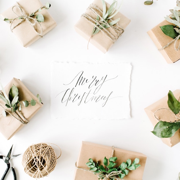 Palabras de caligrafía feliz navidad y marco de arreglo de belleza de cajas artesanales y ramas verdes sobre fondo blanco.