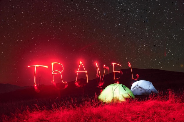 Palavra de viagem de néon vermelho duas tendas iluminadas sob estrelas nas montanhas à noite