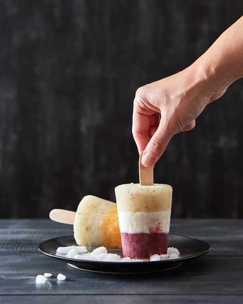 Paleta de hielo de bayas de color en un palo en una placa negra con trozos de hielo sobre un fondo de madera negra con espacio para texto. La mano de la niña toma helado.