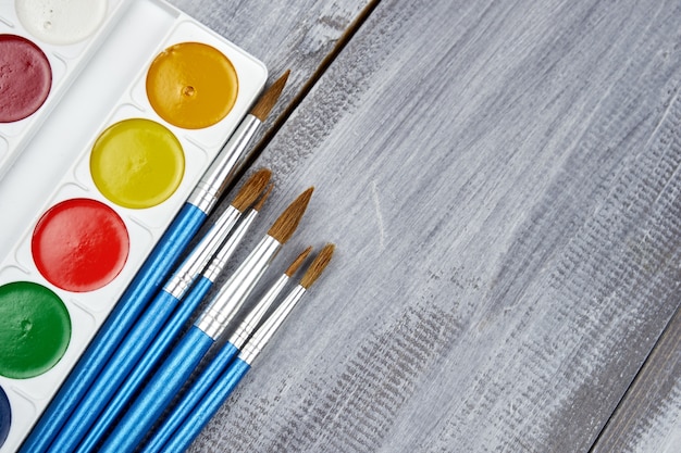 Una paleta de pinturas de acuarela y pinceles de diferentes tamaños, sobre una madera gris.