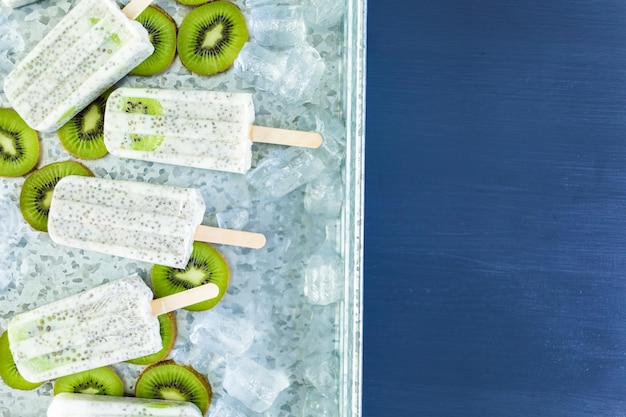 Foto paletas de kiwi, coco y chía con kiwi fresco y hielo en bandeja de metal.