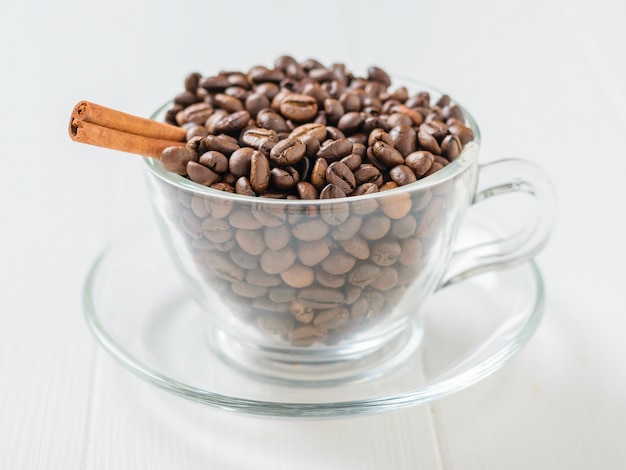 Palillo de canela en granos de café asados en un bol de vidrio en una tabla de madera blanca.