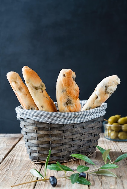 Palitos de pan con aceitunas verdes en la cesta aceite de oliva rama de olivo aceitunas en un tazón sobre una mesa de madera con fondo oscuro