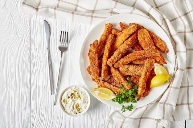 Foto palitos de pescado, filetes de pescado empanizados y fritos servidos en un plato blanco sobre una mesa de madera con salsa tártara y rodajas de limón, vista horizontal desde arriba, espacio libre plano