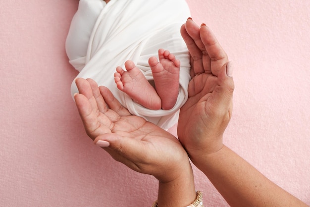 Las palmas de las manos de los padres, el padre y la madre, sostienen las piernas, los pies de un bebé recién nacido en una envoltura blanca sobre un fondo rosa, los pies, los talones y los dedos de los ojos de un niño recién nacido de cerca.