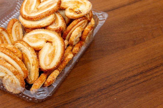 Palmier Kekse - französische Kekse aus Blätterteig (auch Palmblätter, Elefantenohren oder französische Herzen genannt) verpackt in Plastikbox auf Holztisch