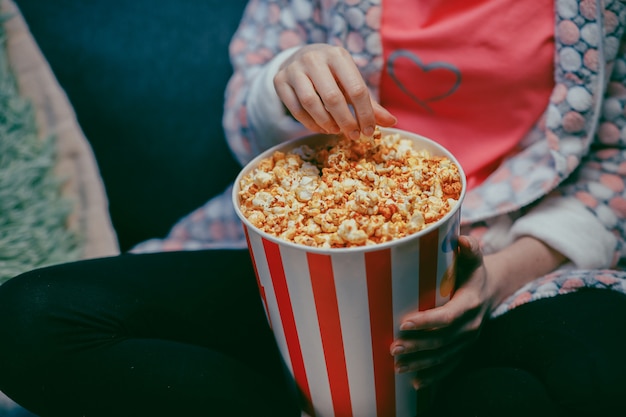 Palomitas de maíz de la cosecha de la mano femenina del primer del cubo de papel. Ciérrese para arriba de la mujer que come palomitas de maíz en el cine. Mano femenina tomando palomitas de maíz en el cubo en el cine.