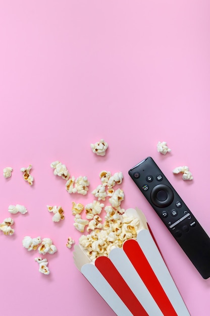 Palomitas de maíz derramadas de un vaso de cartón rayado y un control remoto sobre un fondo rosa con espacio para copiar El concepto de ocio viendo películas muestra deportes Vista vertical