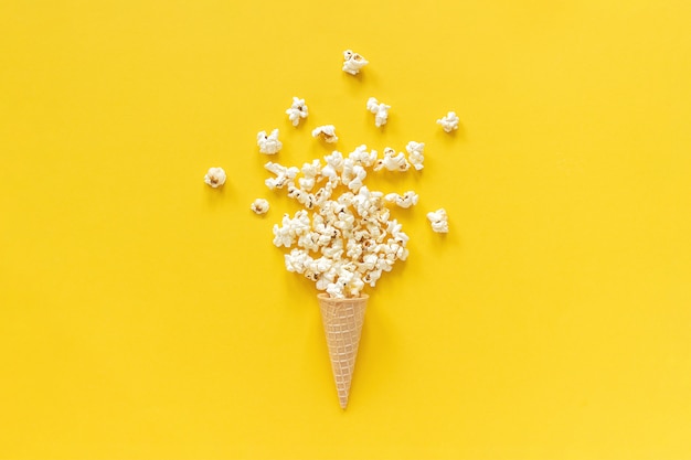 Palomitas de maíz dispersas en cono de la galleta del helado en el fondo de papel amarillo.