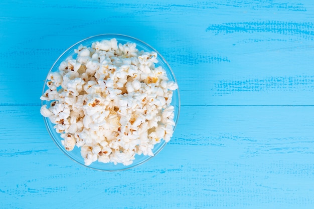 Palomitas de maíz saladas blancas en un recipiente de vidrio sobre una mesa azul. Vista superior con espacio de copia