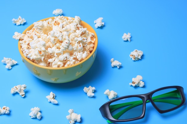 Palomitas de maíz en una taza amarilla sobre un fondo turquesa y junto a unas gafas 3d para ver una película