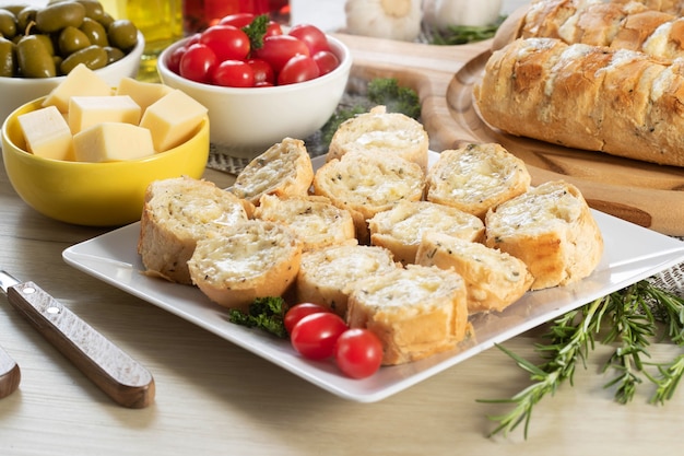 Pan de ajo en plato cuadrado blanco sobre la mesa con queso, romero, aceitunas y tomates cherry.