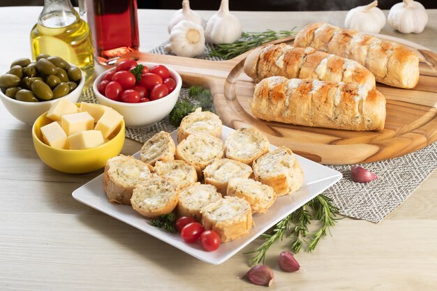 Pan de ajo en plato cuadrado blanco sobre la mesa con queso, romero, aceitunas y tomates cherry.