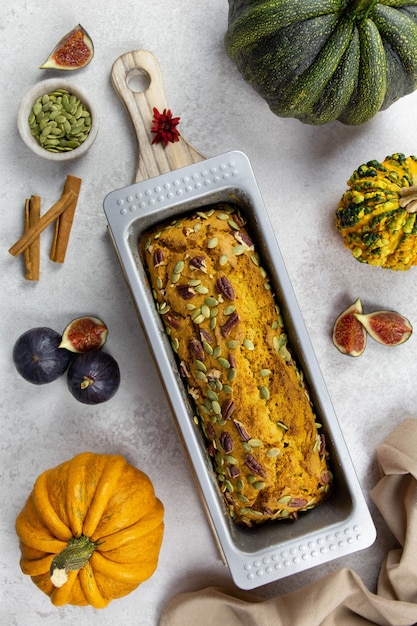 Pan de calabaza, pastel sobre una tabla de cortar de madera con nueces pecanas, semillas de calabaza y especias de canela sobre una superficie gris claro con coloridas calabazas e higos