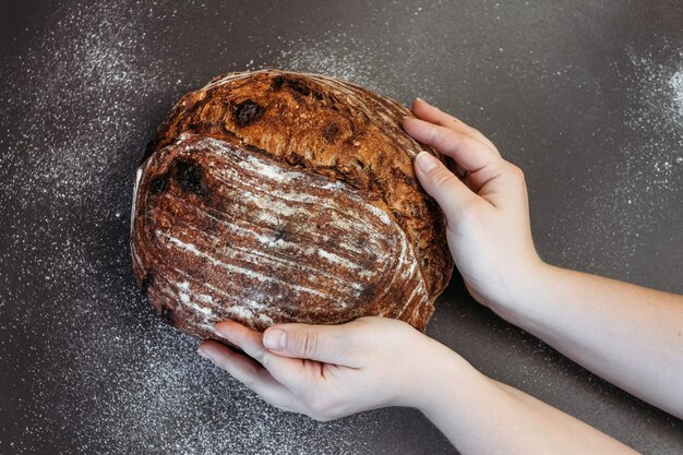 Pan de centeno casero recién horneado, vista superior. Pan con nueces y frutas confitadas. Manos femeninas sostienen pan, fondo oscuro.