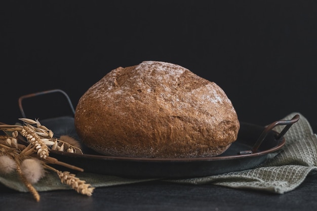 Un pan de centeno redondo en una placa de metal vintage con una servilleta de cocina y orejas yacen sobre un fondo de piedra negra vista lateral de primer planoConcepto de horneado de pan