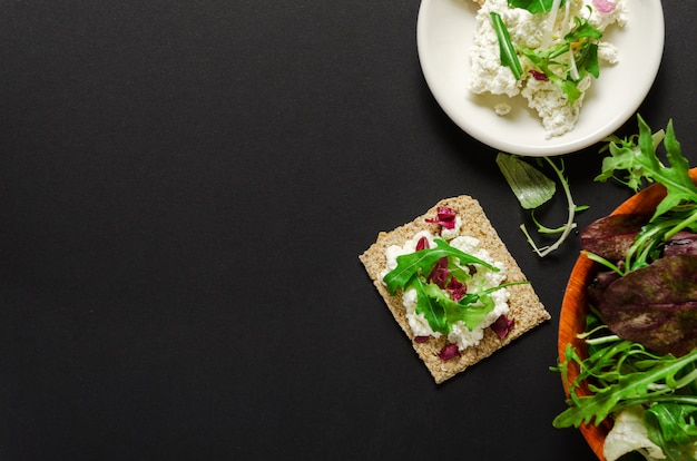 Pan crujiente, queso crema en un plato, mezcla de verduras frescas sobre un fondo negro.