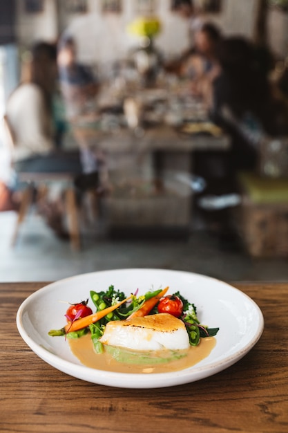 Foto pan filé de bacalhau com tomate, feijão e cenoura em molho de molho. servido em chapa branca na mesa de madeira com borrão pessoas e mesa de jantar