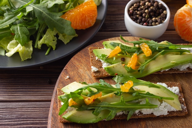 Foto pan integral crujiente con queso, aguacate, hojas frescas y mandarina sobre fondo de madera