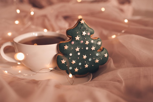 Pan de jengibre en forma de árbol de Navidad con una taza de té negro sobre las luces de Navidad de cerca. Temporada de vacaciones de invierno.